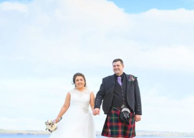 a bride and groom smile as they walk along the beach at their stornoway wedding