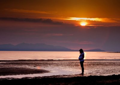 a pregnant mother is silhouetted against the sun on a maternity session on troon beach