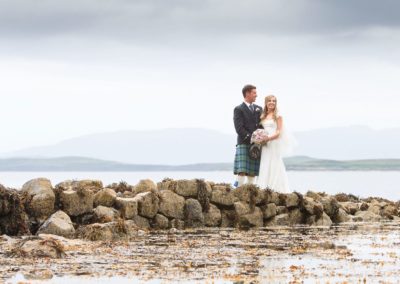 a bride and groom stand on rocks by the waters edge in a scottish wedding photograph from the isle of skye