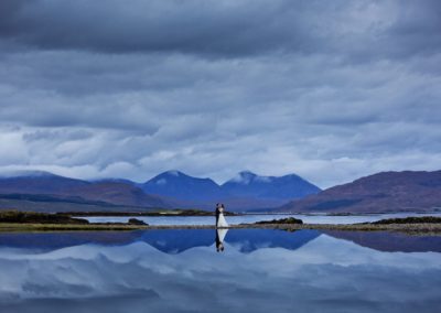 a bride and groom are reflected off the water along with the cuilin hills on the isle of skye